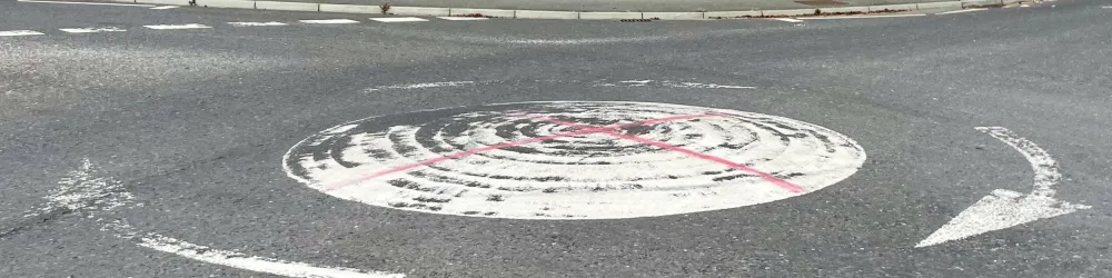 A mini-roundabout with worn white paint has two red lines spray-painted on to form a rudimentary St George's Cross