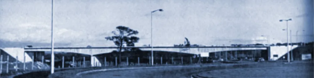 Black and white photograph of a roundabout forming part of a motorway interchange, with streetlights and trees in the foreground. A long arched motorway bridge stretches across the background from left to right