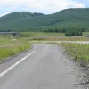 The carriageway edge line gives away the road's former alignment as it the burned out remains of the line head straight ahead while the cycle lane has been curved to provide a perpendicular junction with the B7076. The original A74 continued straight over the path of the motorway and took a slight left turn as it did so.