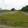 Further north still, the road runs uphill from its modern counterpart, with the cutting made for both carriageways standing out quite obviously. The cutting for the former carriageway stands out quite clearly here. Yet more of the two traffic signs are visible. A layby has remained on the former northbound carriageway, providing a resting point for cyclists. Over to the right, the M74 can be seen sweeping over the moor.